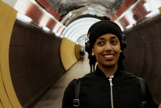 En leende person med hörlurar och svart mössa går genom en upplyst gångtunnel. Bakgrunden visar de runda tunnelväggarna och några personer längre bort.