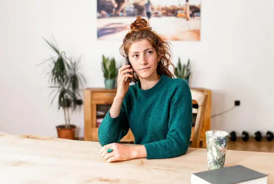 En kvinna med uppsatt hår, lite slarvig uppsättning, ringer ett samtal vid köksbordet.