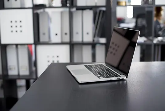 En laptop stående på ett skrivbord med hyllor i bakgrunden.