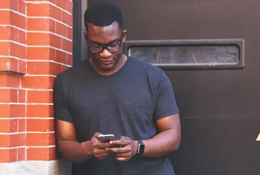Man i en mörkblå t-shirt, ståendes utan för en ytterdörr och surfar på mobilen.