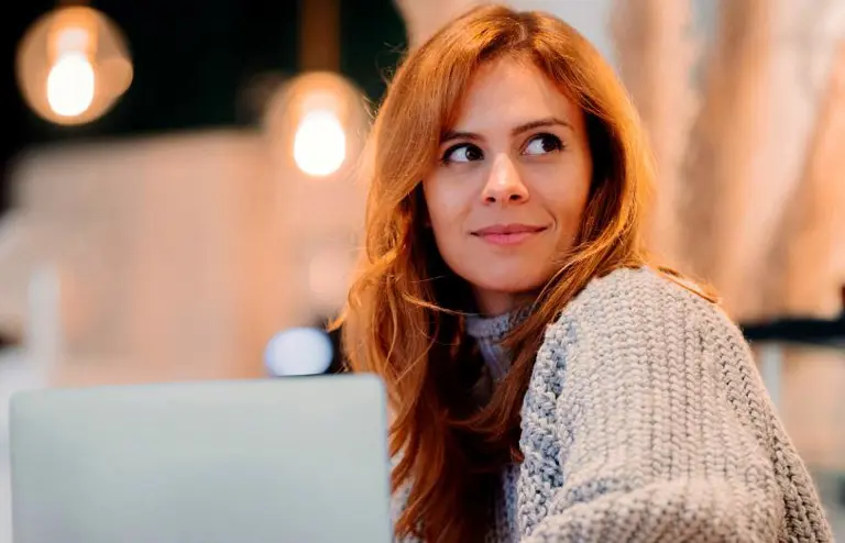 En kvinna med långt rött hår sitter vid en bärbar dator och tittar leende åt sidan. Bakom henne syns mjukt ljus från hängande lampor i en modern, mysig miljö.